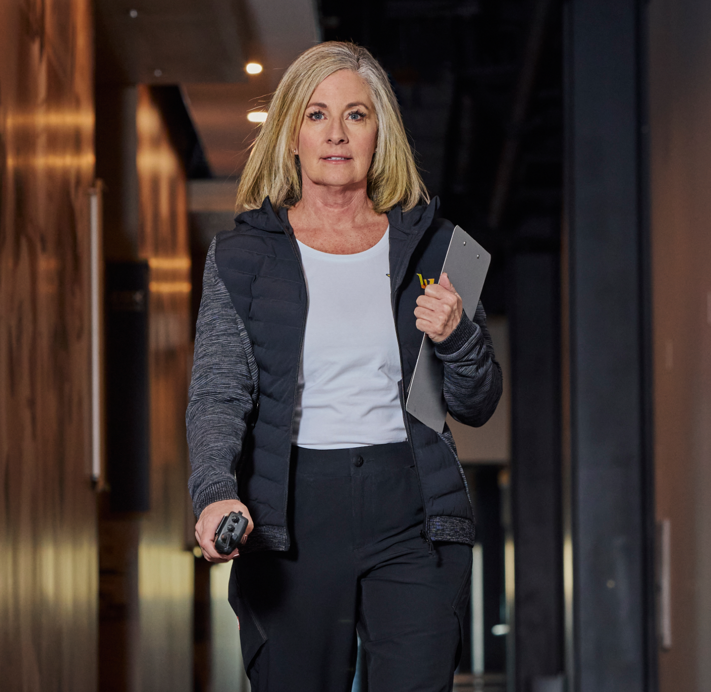 A woman with shoulder-length blonde hair walks confidently down a hallway, holding a clipboard and a remote control. She wears black pants, a white shirt, and a dark vest. The setting appears to be an office or modern building.