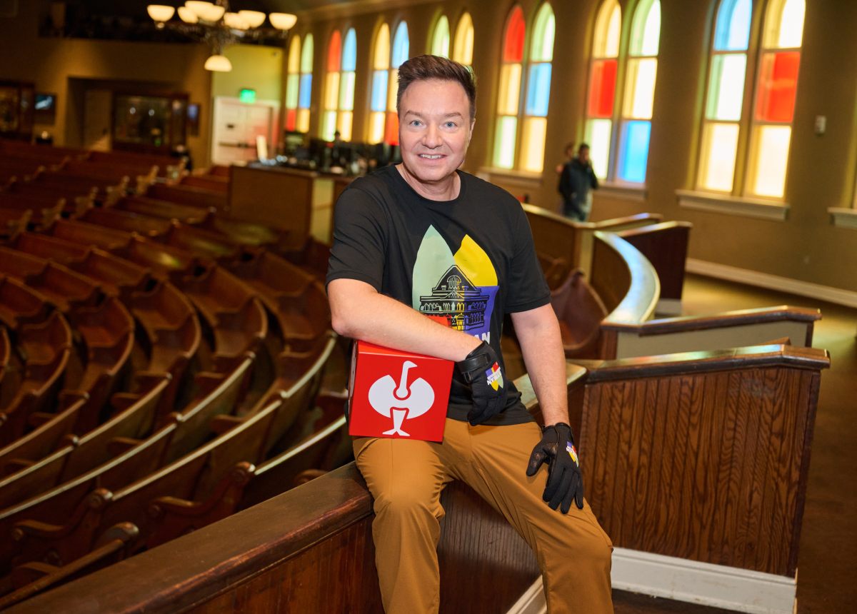 A smiling person in a colorful shirt and brown pants sits on a wooden bench in an empty theater, holding a red box with a white muscle arm graphic, with stained glass windows in the background.