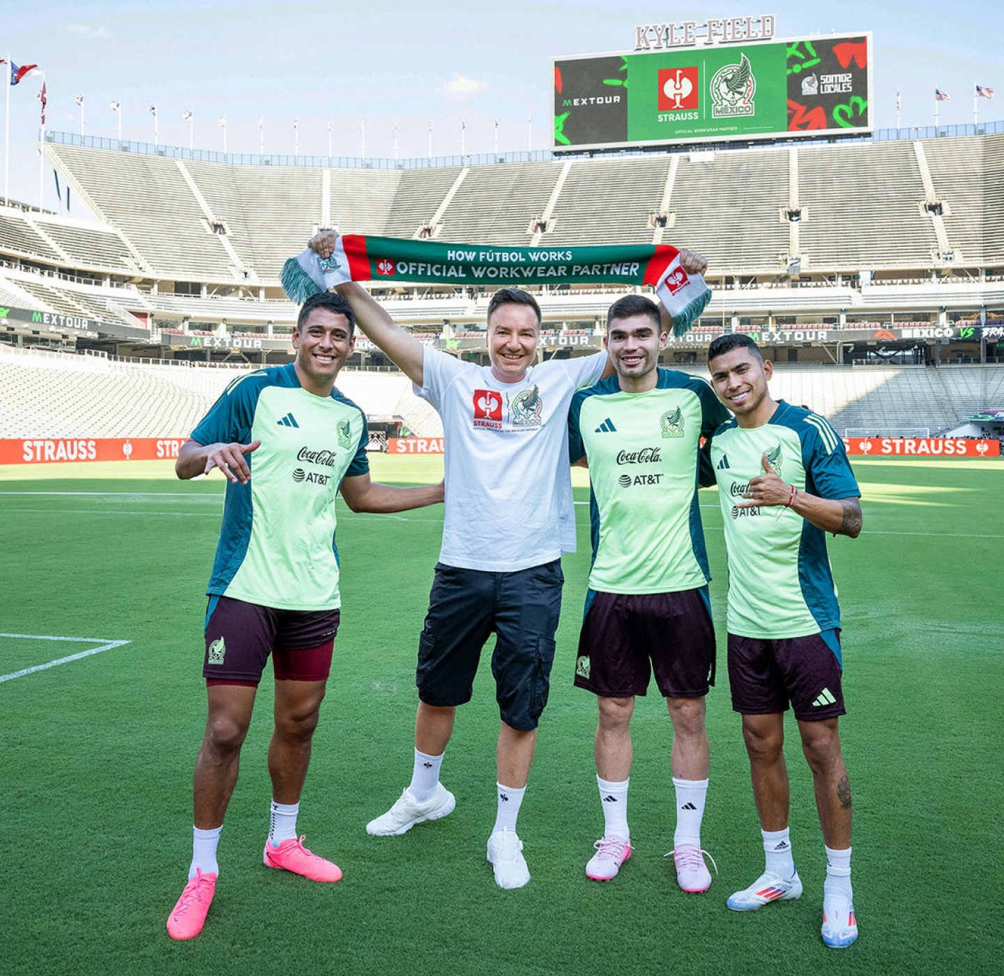 Four men stand on a soccer field smiling, with one holding a green and white scarf above his head. All wear sports attire, and an empty stadium is visible in the background.