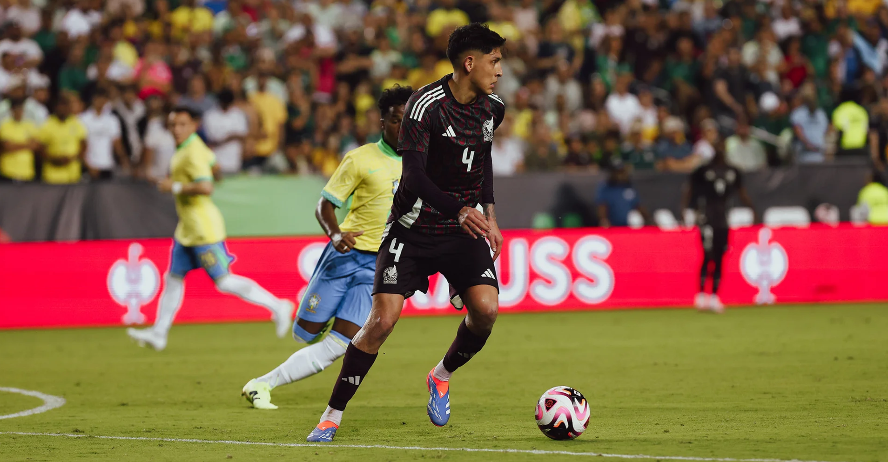 A soccer player in a black uniform controls the ball while a player in a yellow jersey runs behind him on a green field, with a crowd watching from the stands.