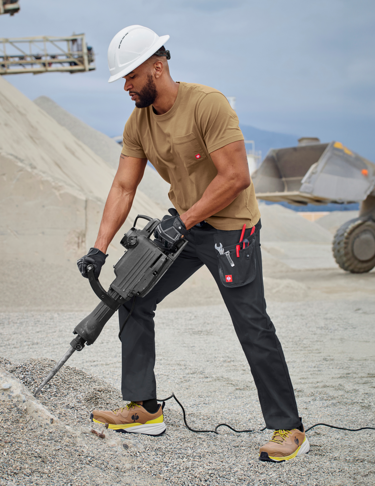 Man in a brown shirt and black pants using a jackhammer on a construction site