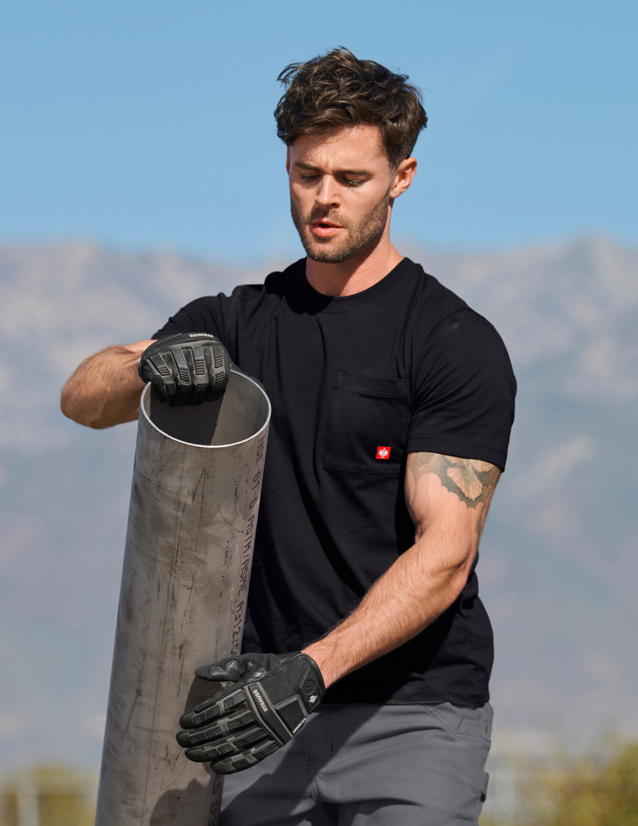 Man in a black shirt holding a cylindrical metal object outdoors with mountains in the background