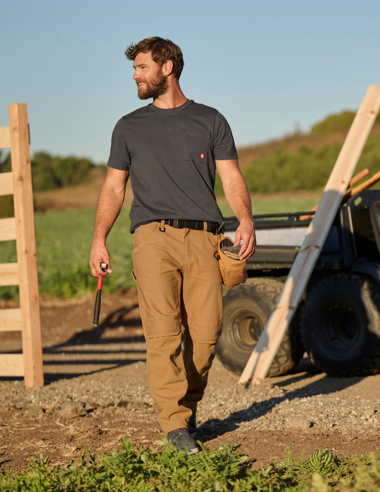 Man in a field wearing a dark shirt and brown pants, holding tools.