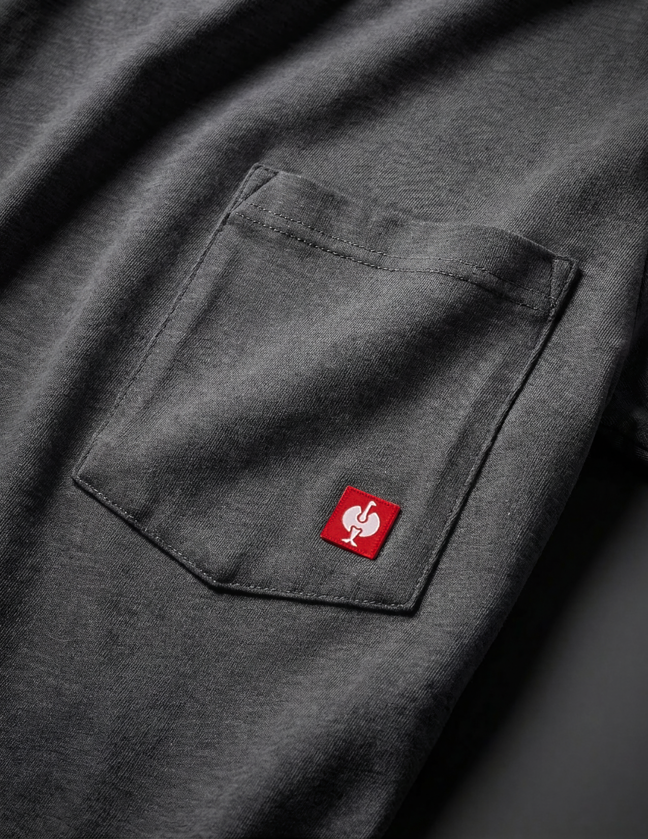 Dark gray shirt with a red logo on the chest pocket against a dark background