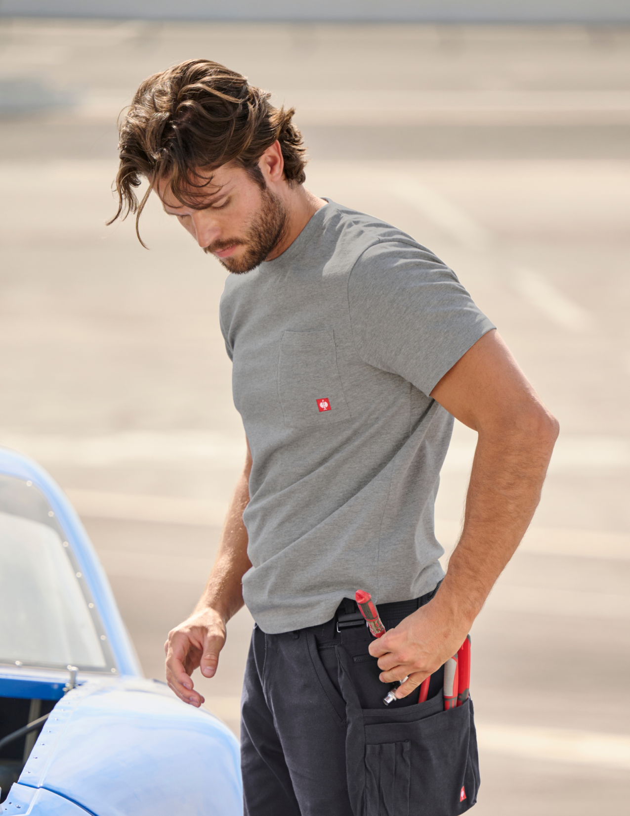 Man in gray t-shirt and dark pants holding a tool near a vehicle.