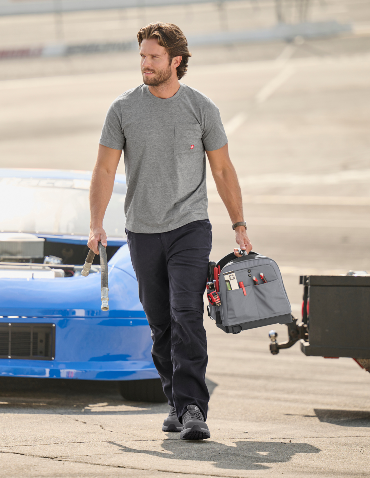 Man in a grey shirt walking with a tool bag near a blue car on a track