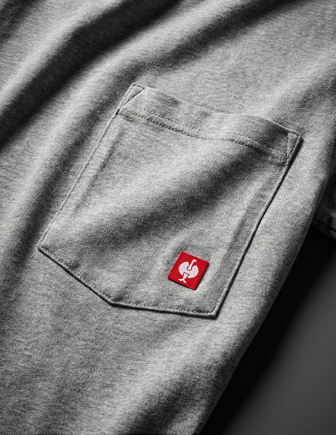 Gray shirt with a red logo on the chest pocket against a dark background