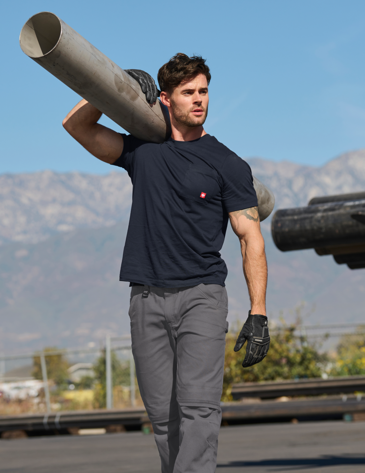 Man in a dark blue T-Shirt carrying a large cylindrical object outdoors with mountains in the background