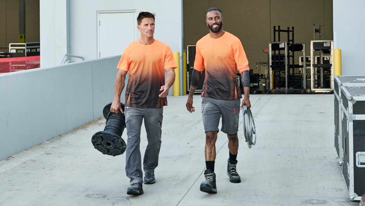 Two men walking in a warehouse corridor wearing matching orange-to-black gradient athletic t-shirts and gray shorts, with one carrying a large black reel and the other holding a coiled cable.