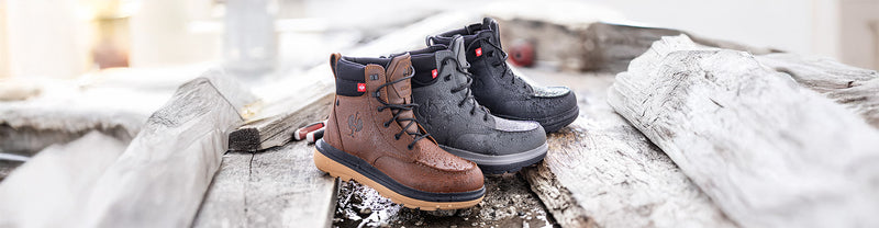 Three pairs of boots, one brown, one black and one gray, on a wooden surface with a blurred background.