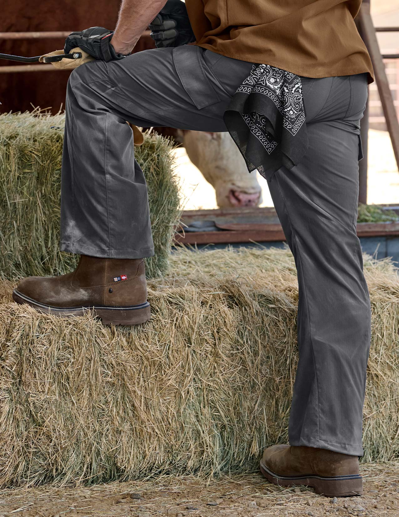 Person standing on a hay bale wearing brown boots, gray pants, and a bandana.