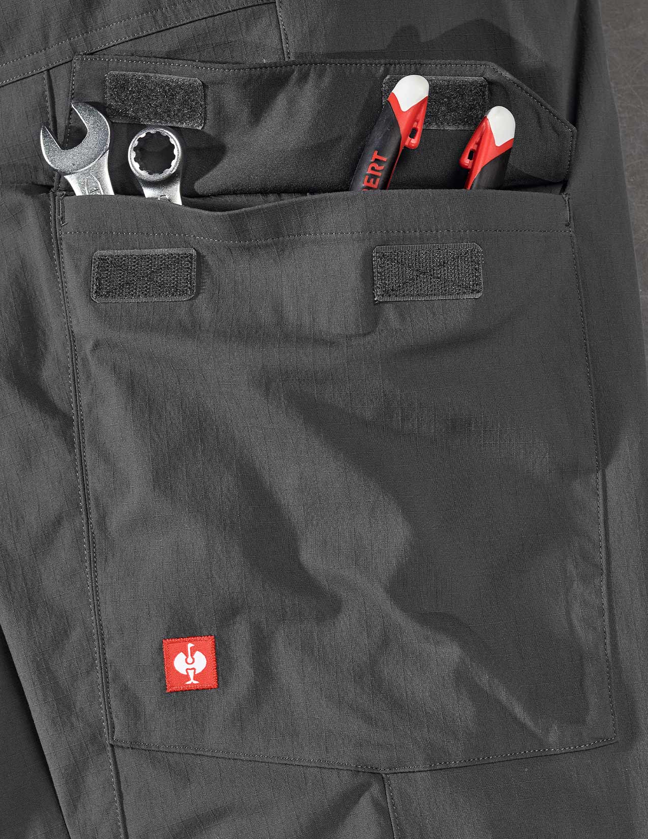 Grey work pants with tools in a pocket and a visible brand logo.