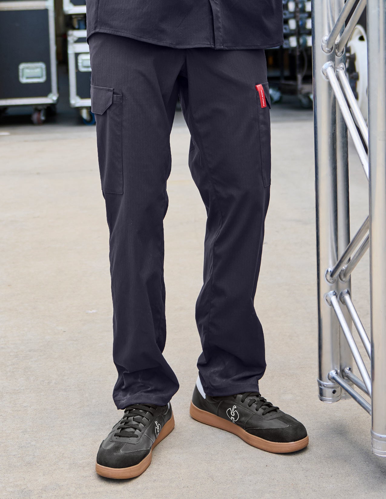 Person wearing dark blue pants and black shoes standing on a concrete surface with equipment in the background