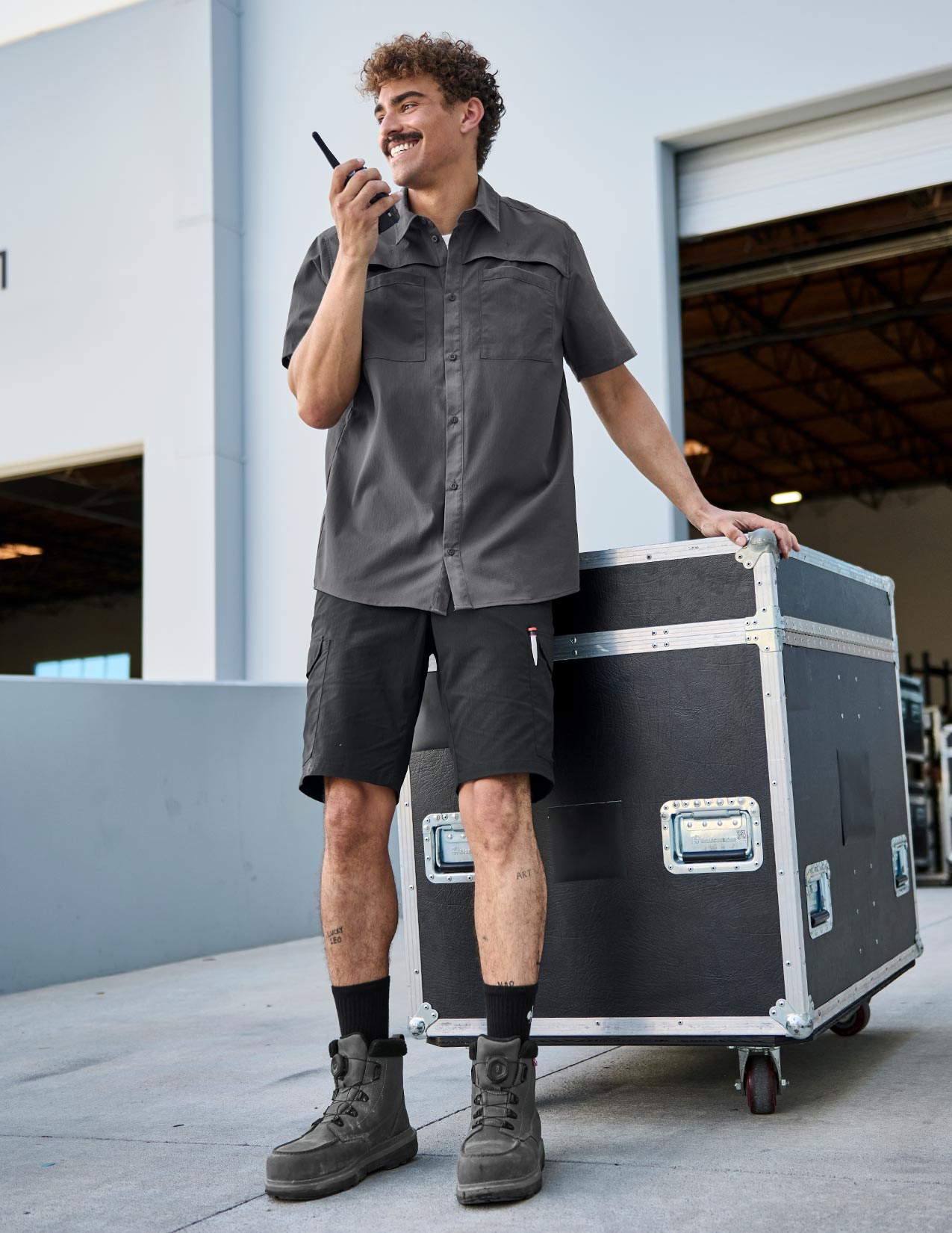 Man standing next to a large black case, holding a walkie-talkie in an outdoor setting.