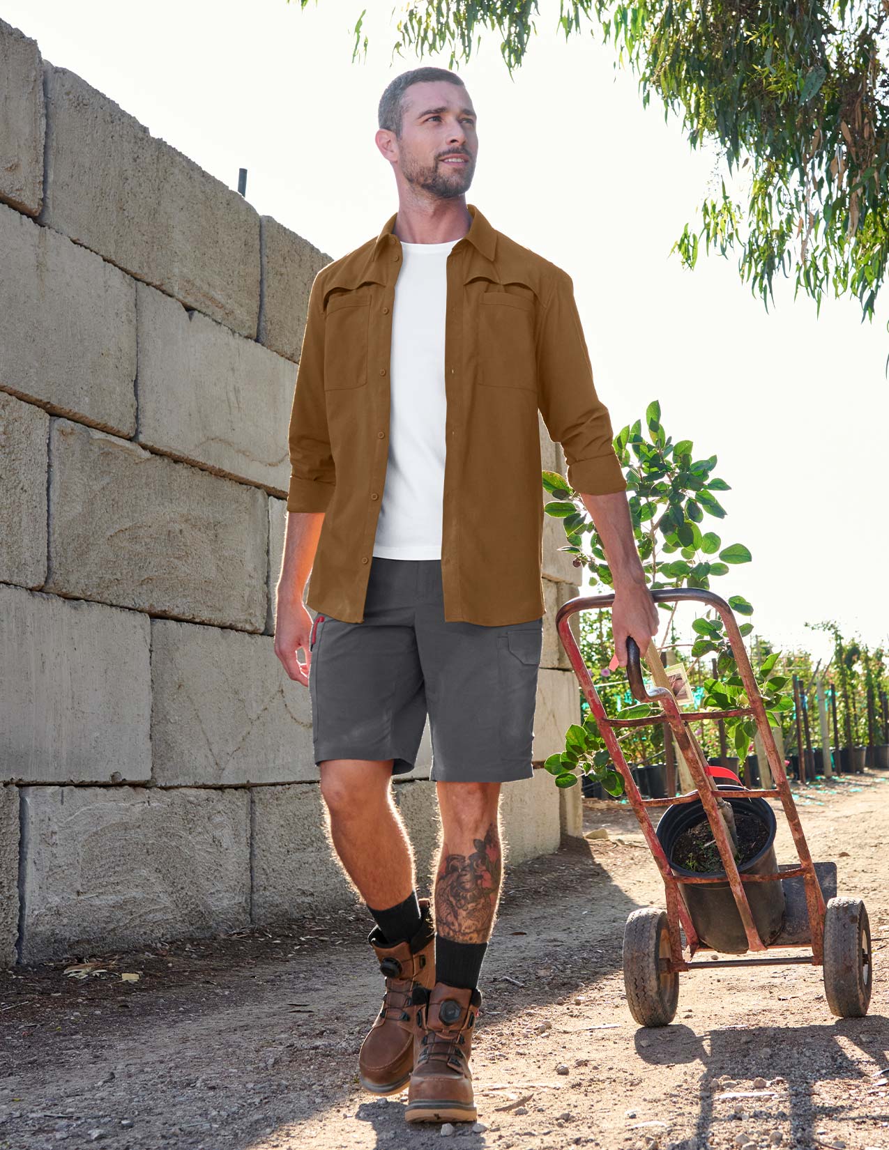 Man in brown jacket and gray shorts walking with a cart outdoors.