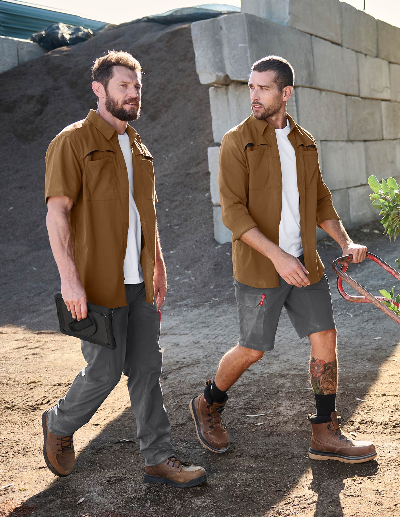 Two men walking outdoors in a casual setting with a stone wall and dirt ground.