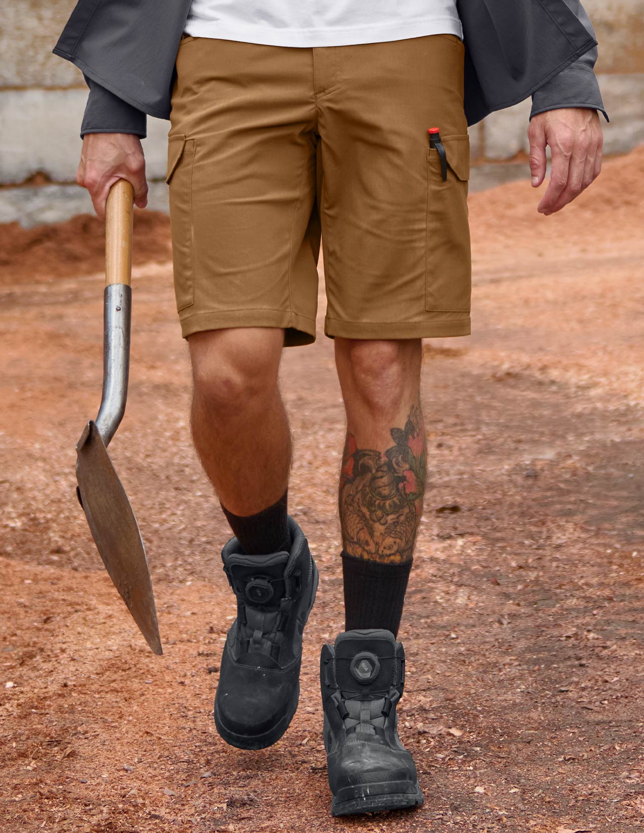Person wearing brown shorts and black boots holding a shovel on a dirt ground.
