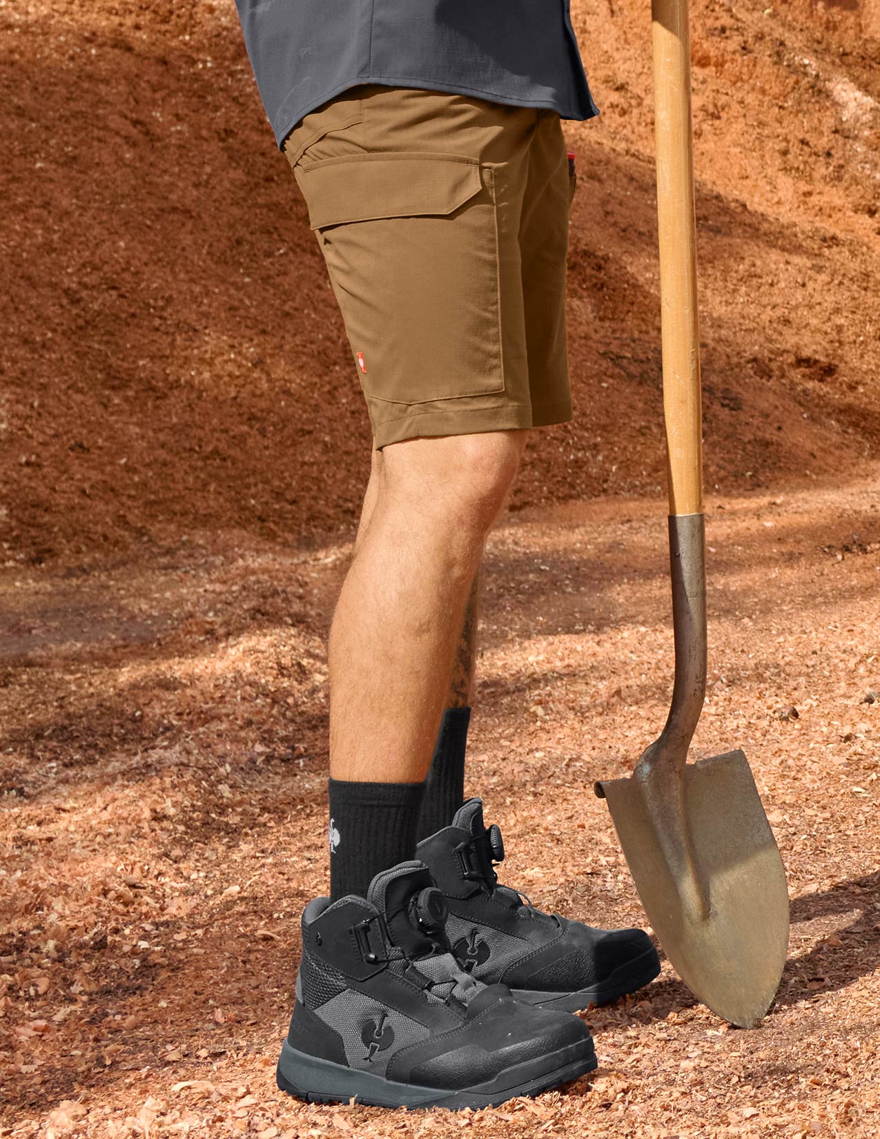 Person wearing brown shorts, black shoes, and black socks holding a shovel on a dirt background