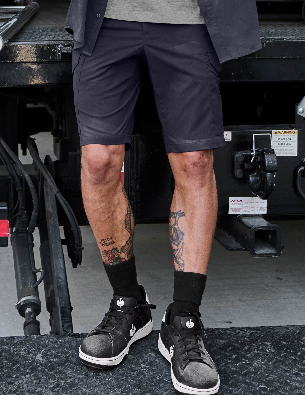 Person wearing navy shorts and black sneakers with visible tattoos on legs, standing in front of a vehicle.