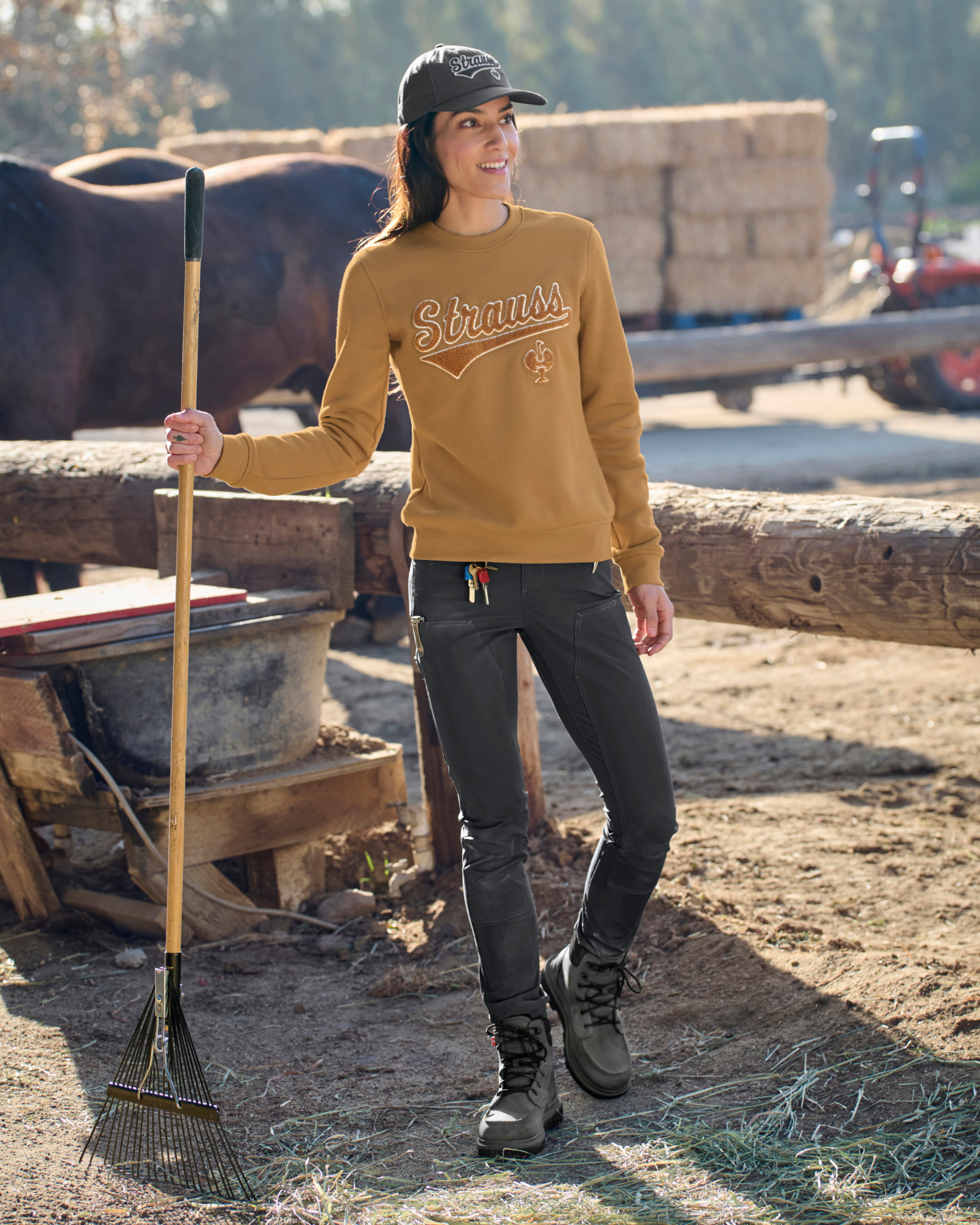Woman in a yellow sweater with 'STRAUSS' logo holding a rake on a farm.