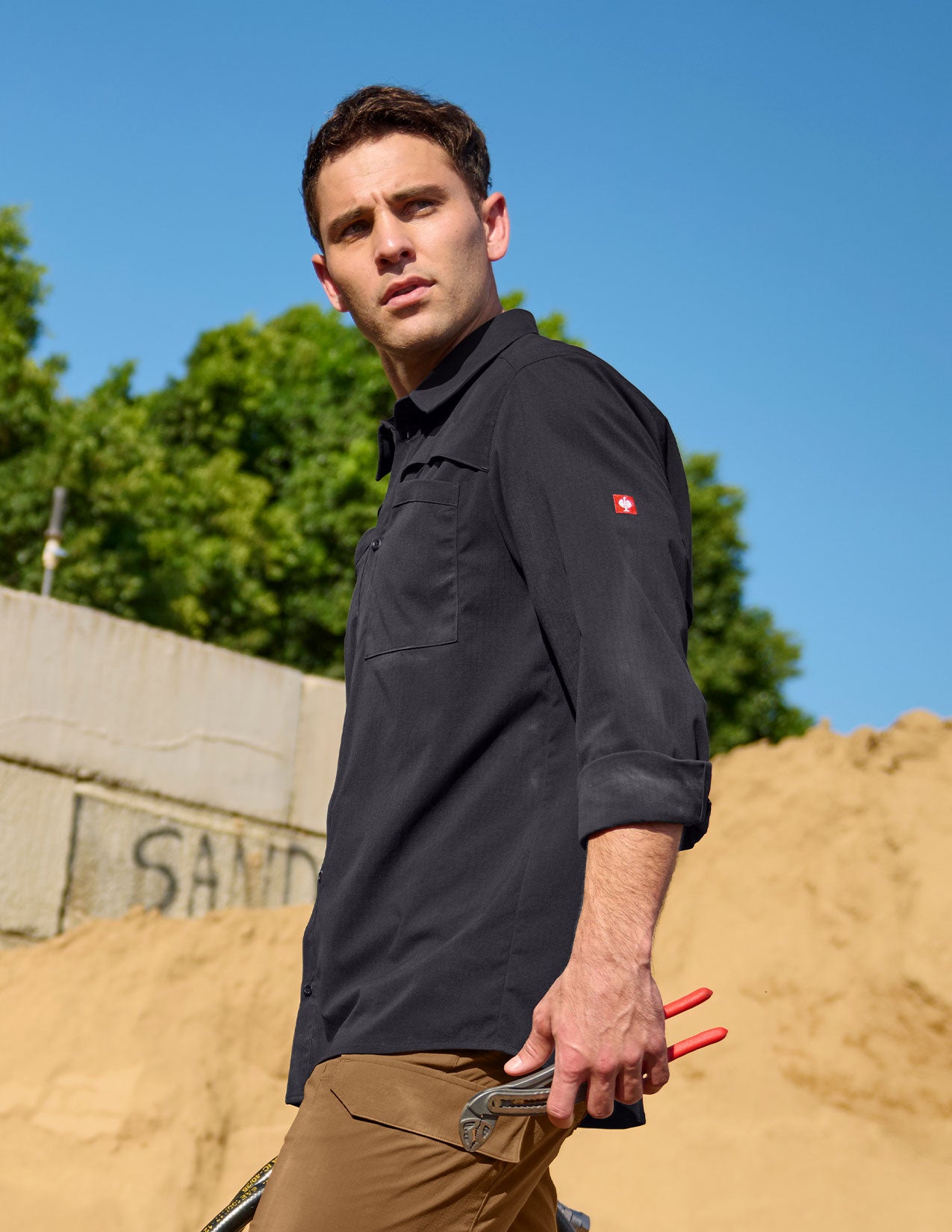 Man wearing a black shirt with a red logo outdoors against a blue sky and sand dunes.