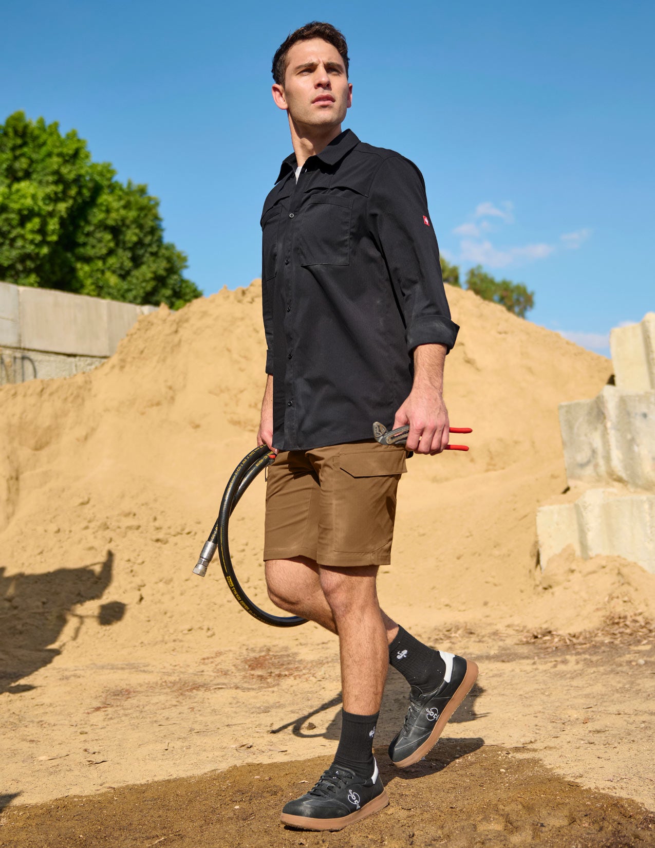 Man in black shirt and brown shorts holding tools against a sandy background