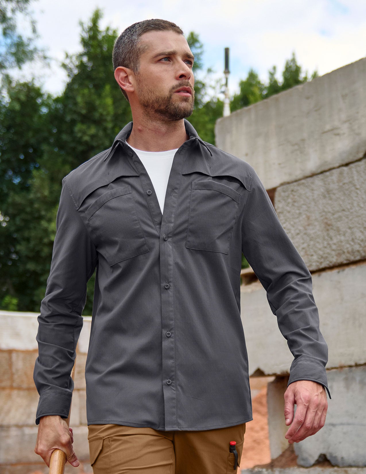 Man wearing a gray shirt outdoors with trees and concrete wall in the background