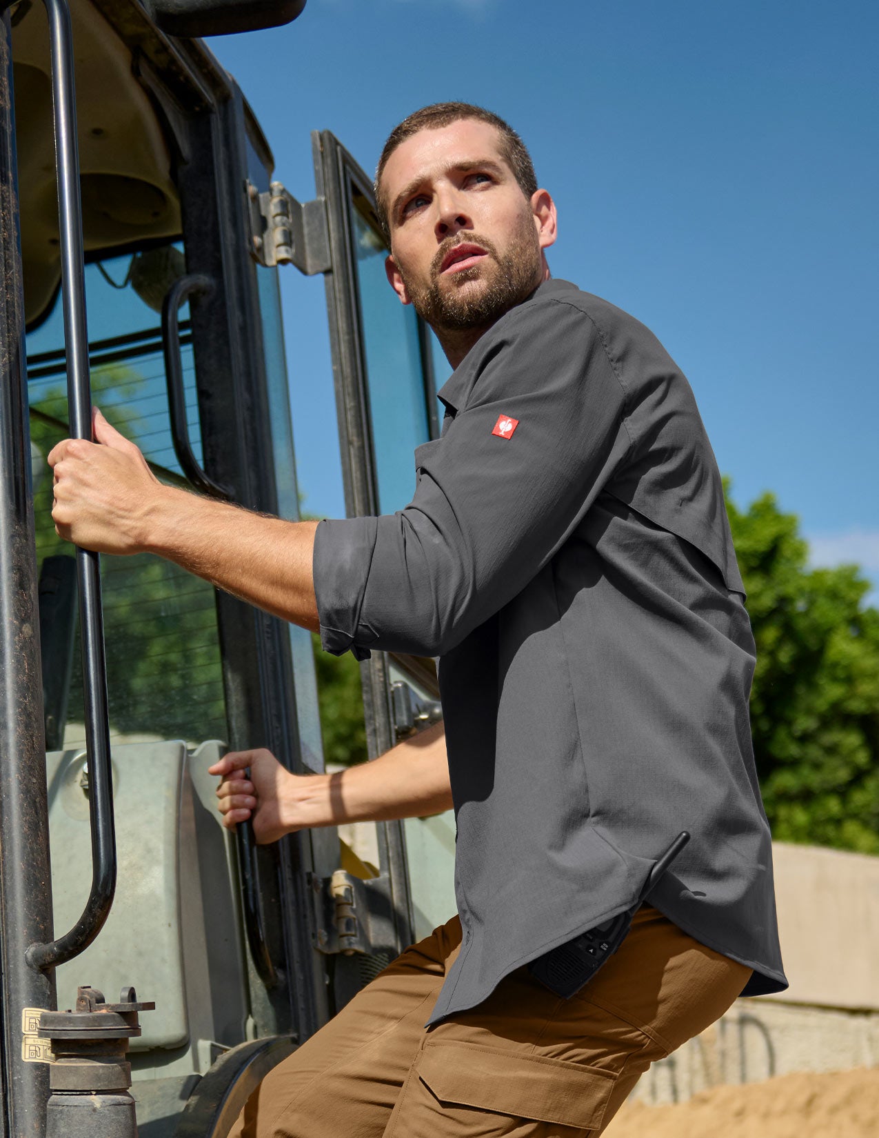 Man in a gray shirt and brown pants standing next to a large piece of machinery with a clear blue sky in the background.