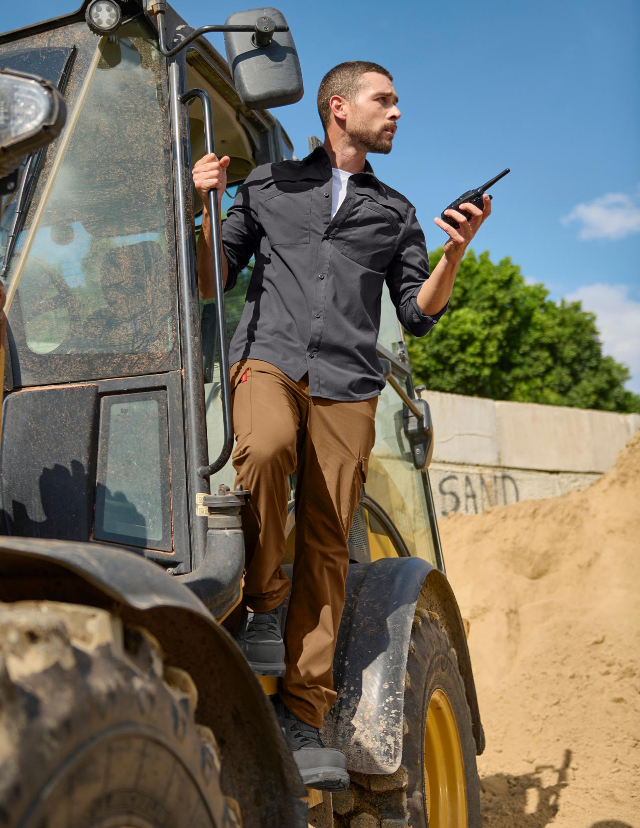 Man standing next to a large vehicle holding a walkie-talkie, with a clear sky and greenery in the background.