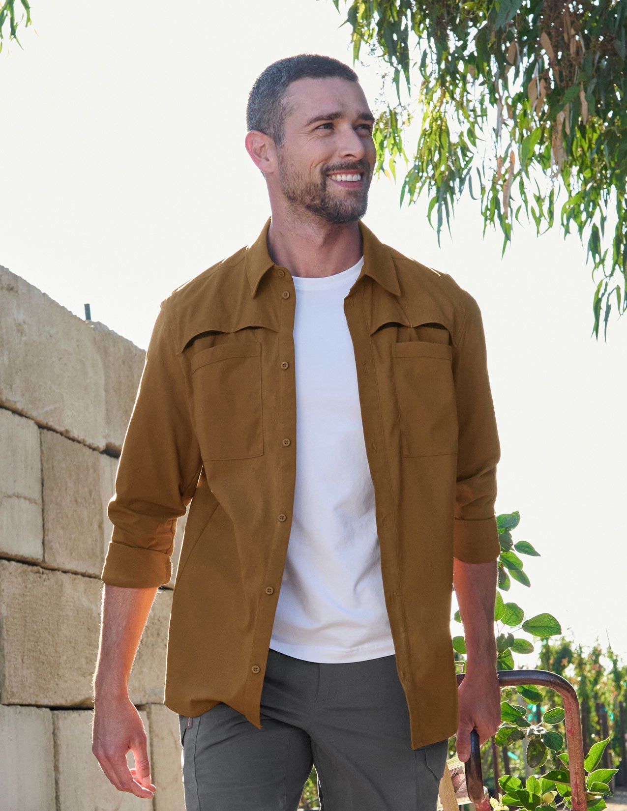 Man wearing a brown jacket over a white shirt, standing outdoors with greenery in the background.