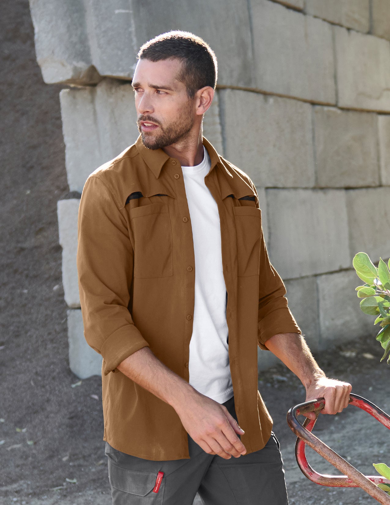 Man wearing a brown work shirt and white shirt standing against a stone wall.