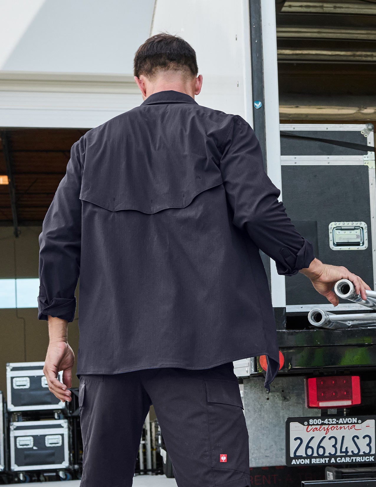 Person wearing a dark blue work shirt and pants standing next to a vehicle with equipment, shown from the back