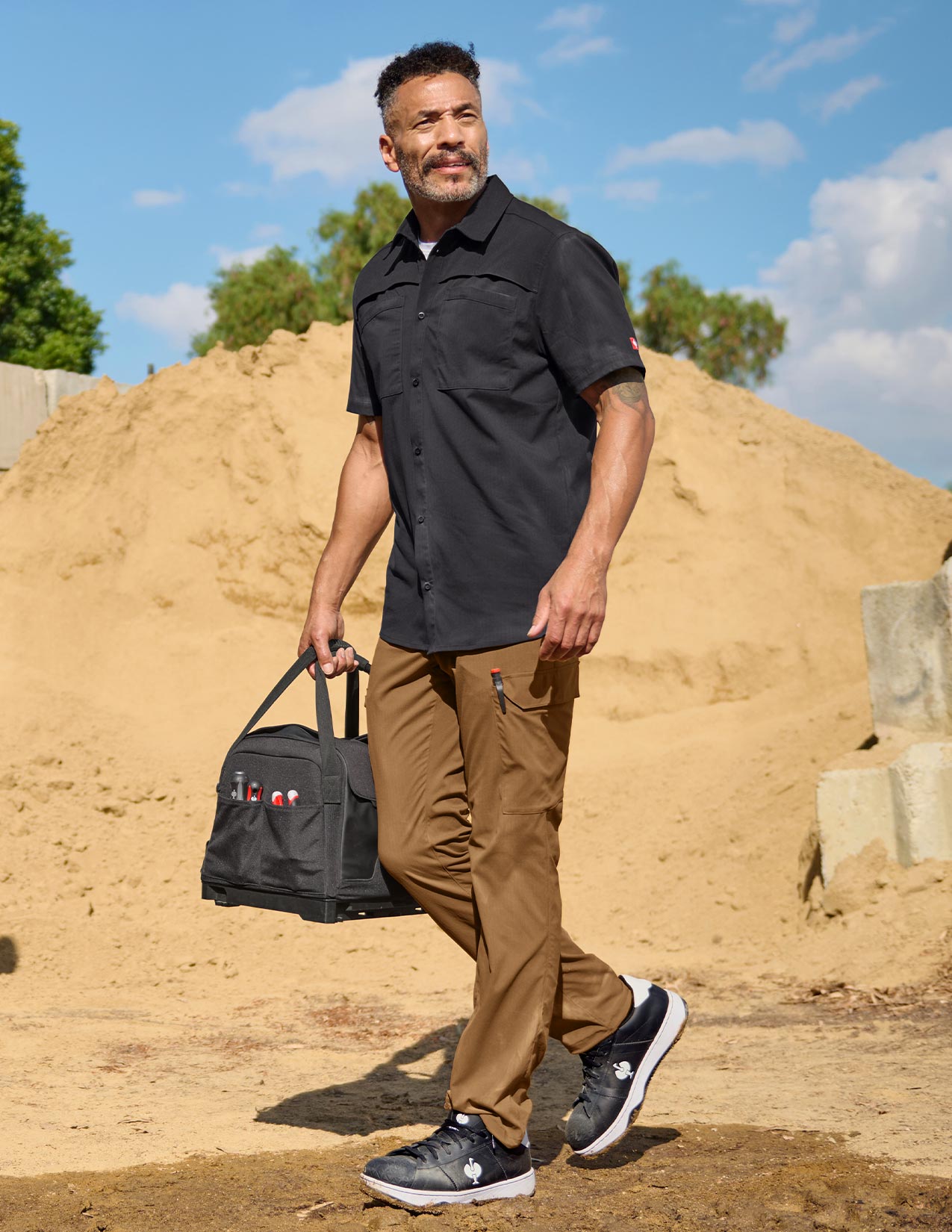 Man in a black short sleeved Work Shirt and brown pants standing outdoors.
