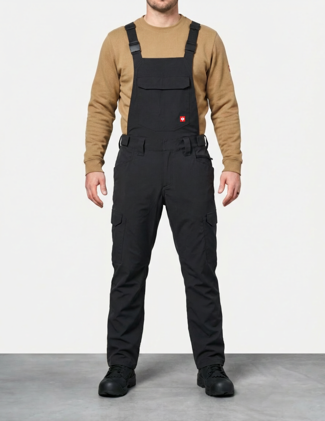 Person wearing black bib overalls with a beige sweater on a white background