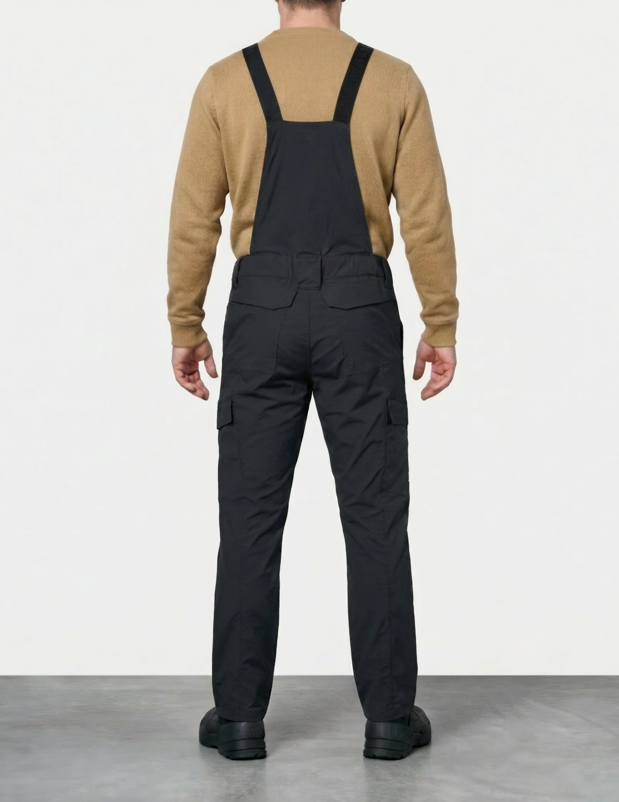 Person wearing black overalls and a beige sweater on a white background
