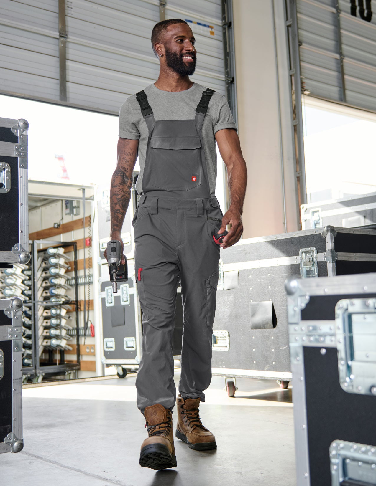 A man wearing gray cargo work bibs and holding a drill in his hand