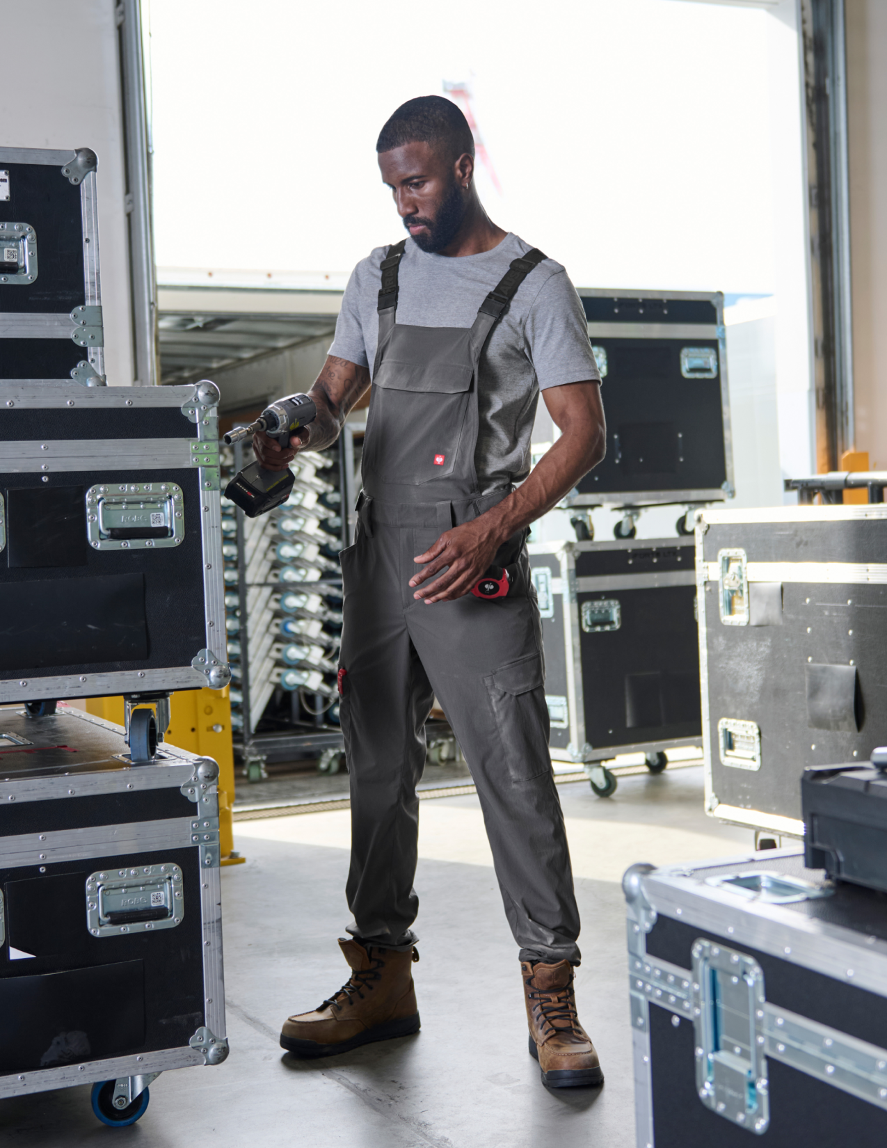 A man wearing gray cargo work bibs and holding a drill in is hand