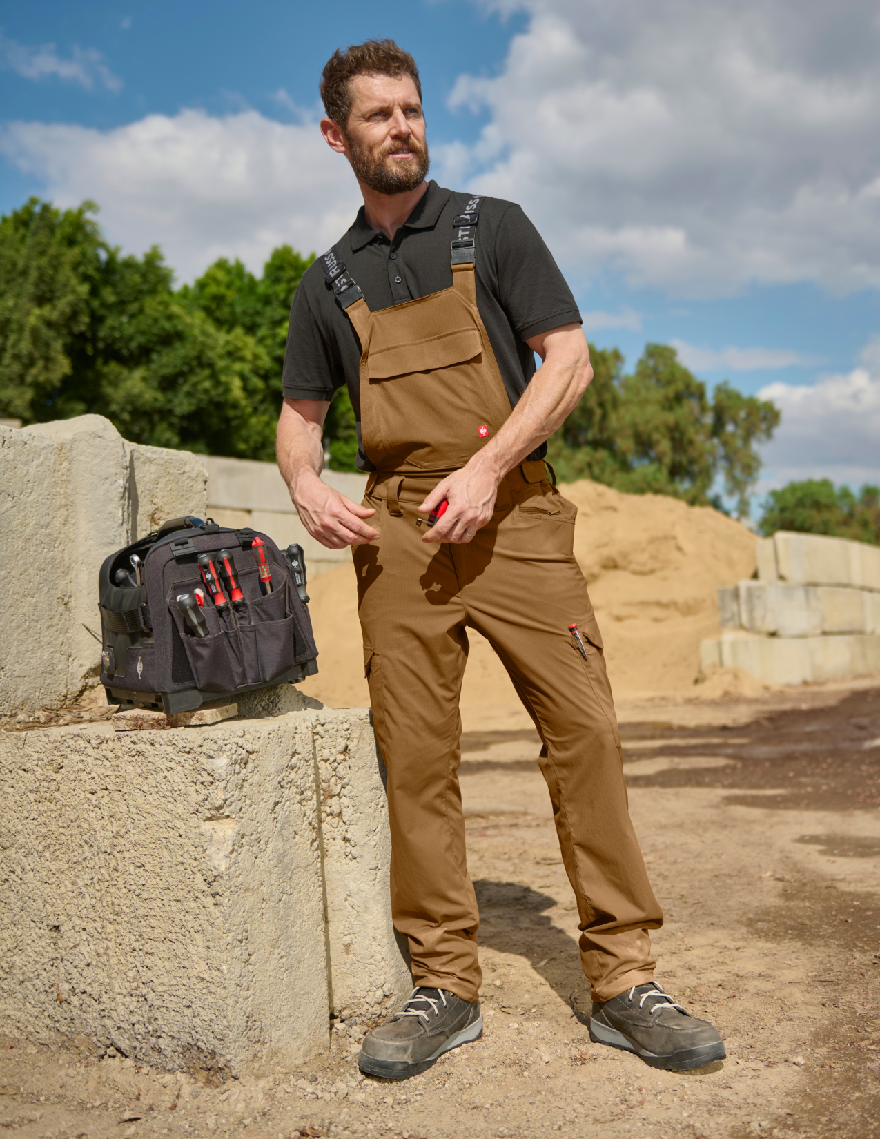 Man wearing brown overalls standing outdoors with a tool bag.