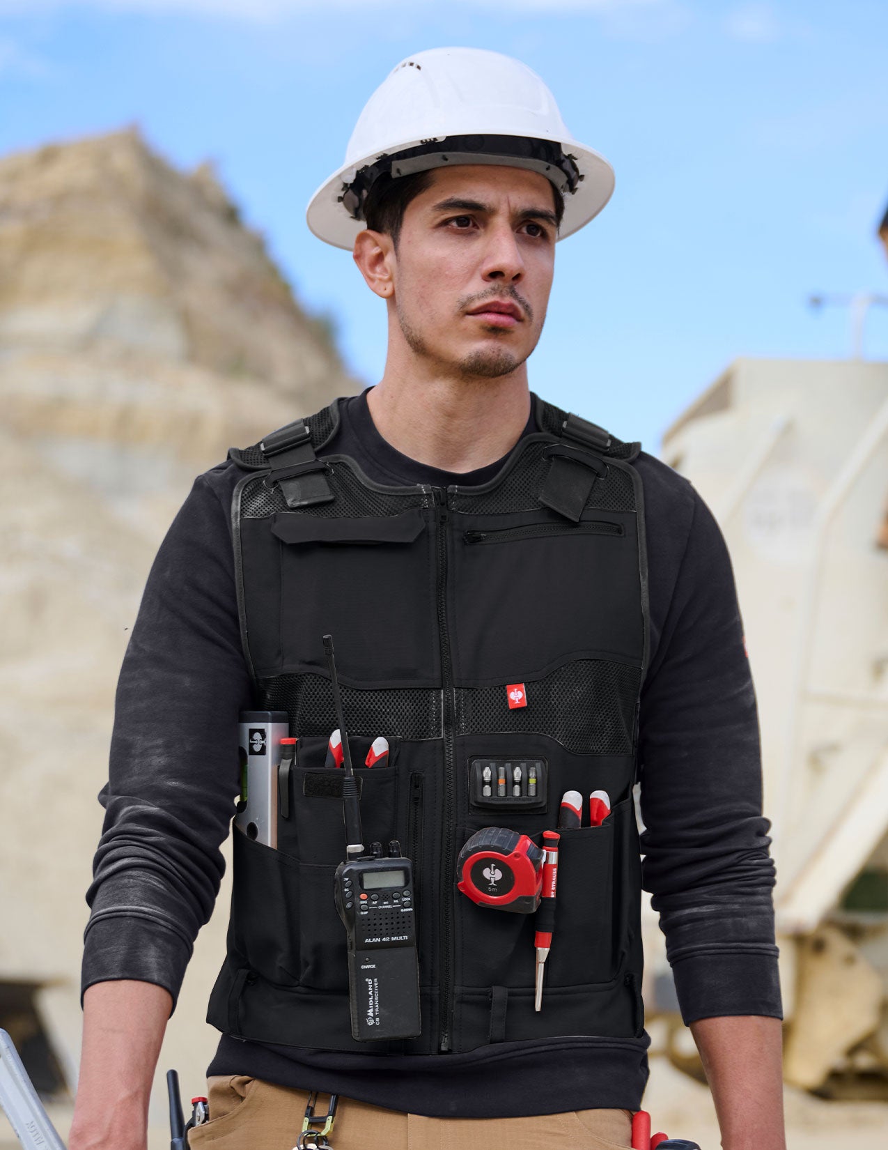 Person wearing a black utility vest with tools, standing outdoors.