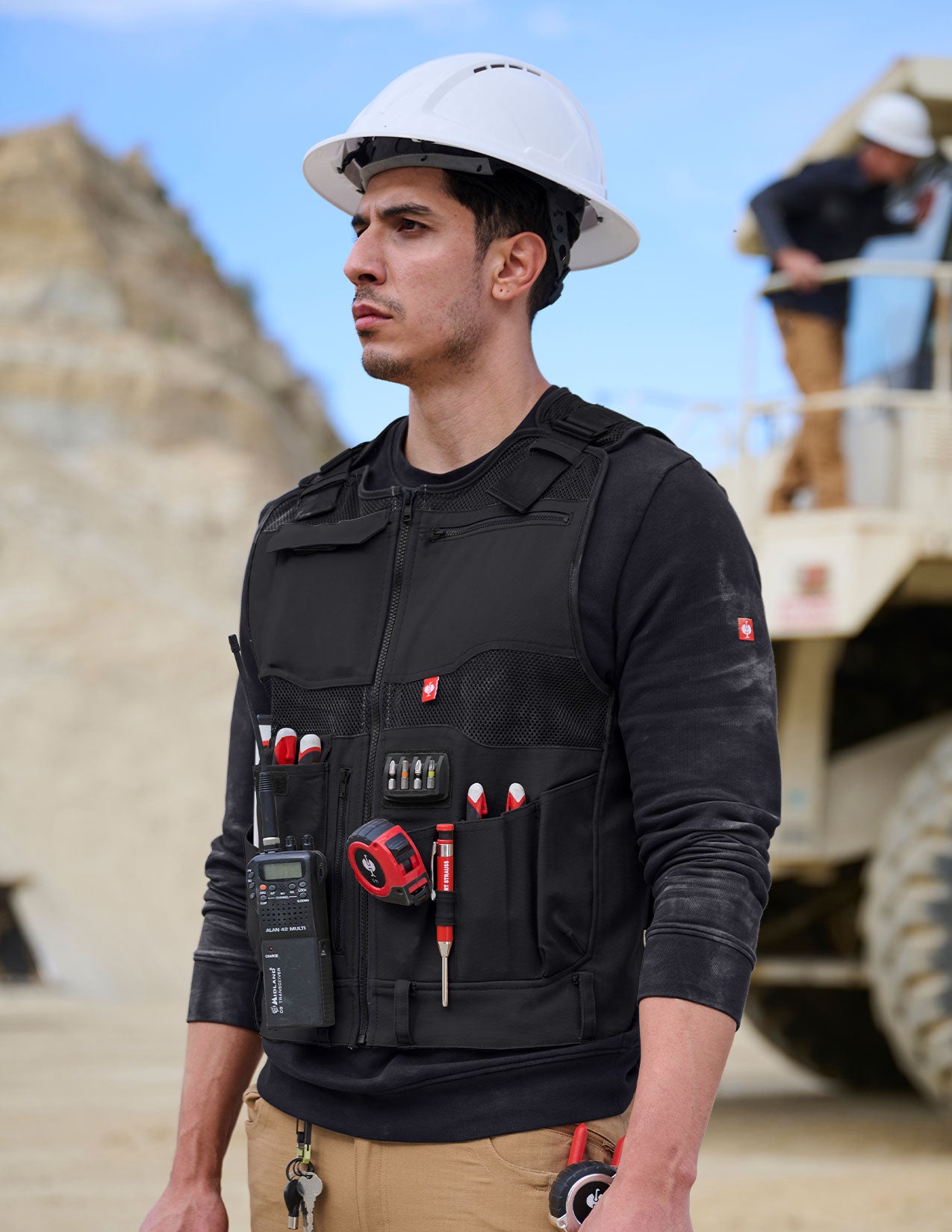 Person wearing a black tool vest with tools and a hard hat in an outdoor construction setting.