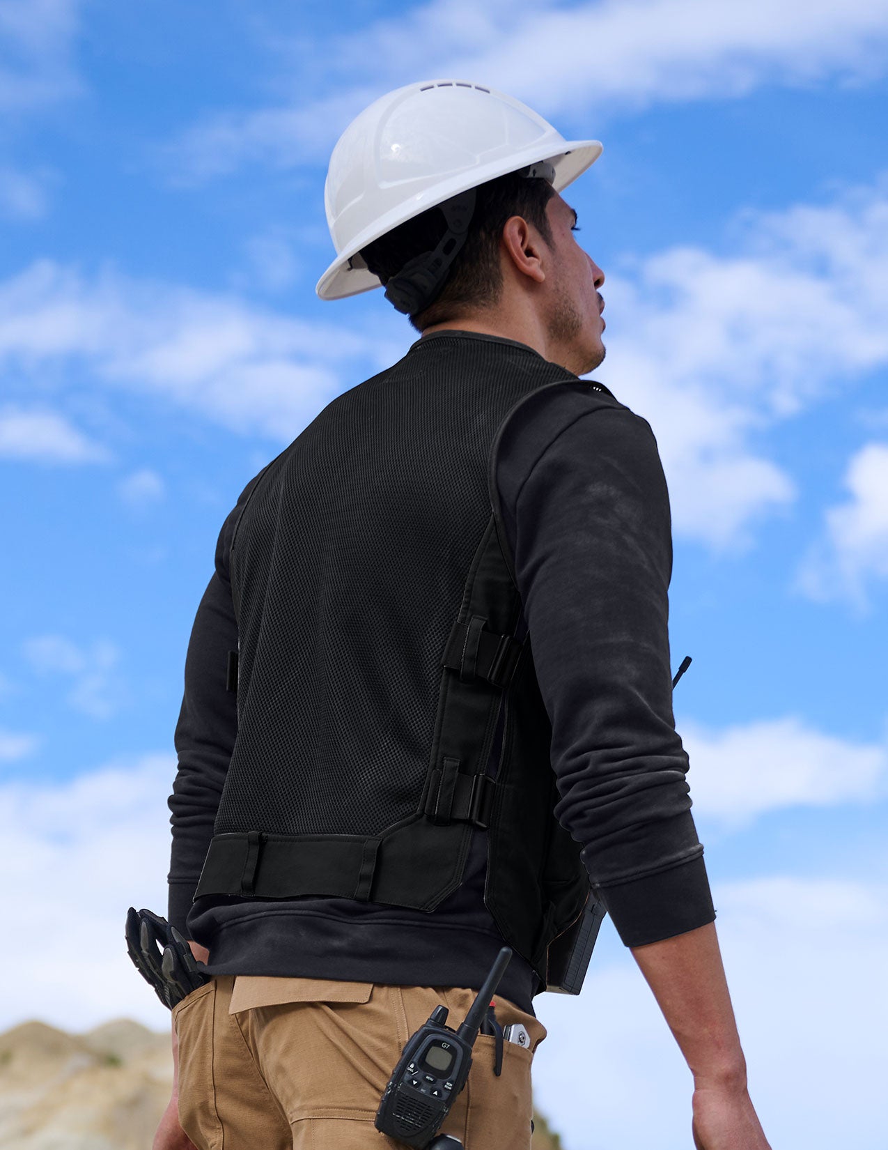Person wearing a black vest, beige pants, and a white hard hat against a blue sky.