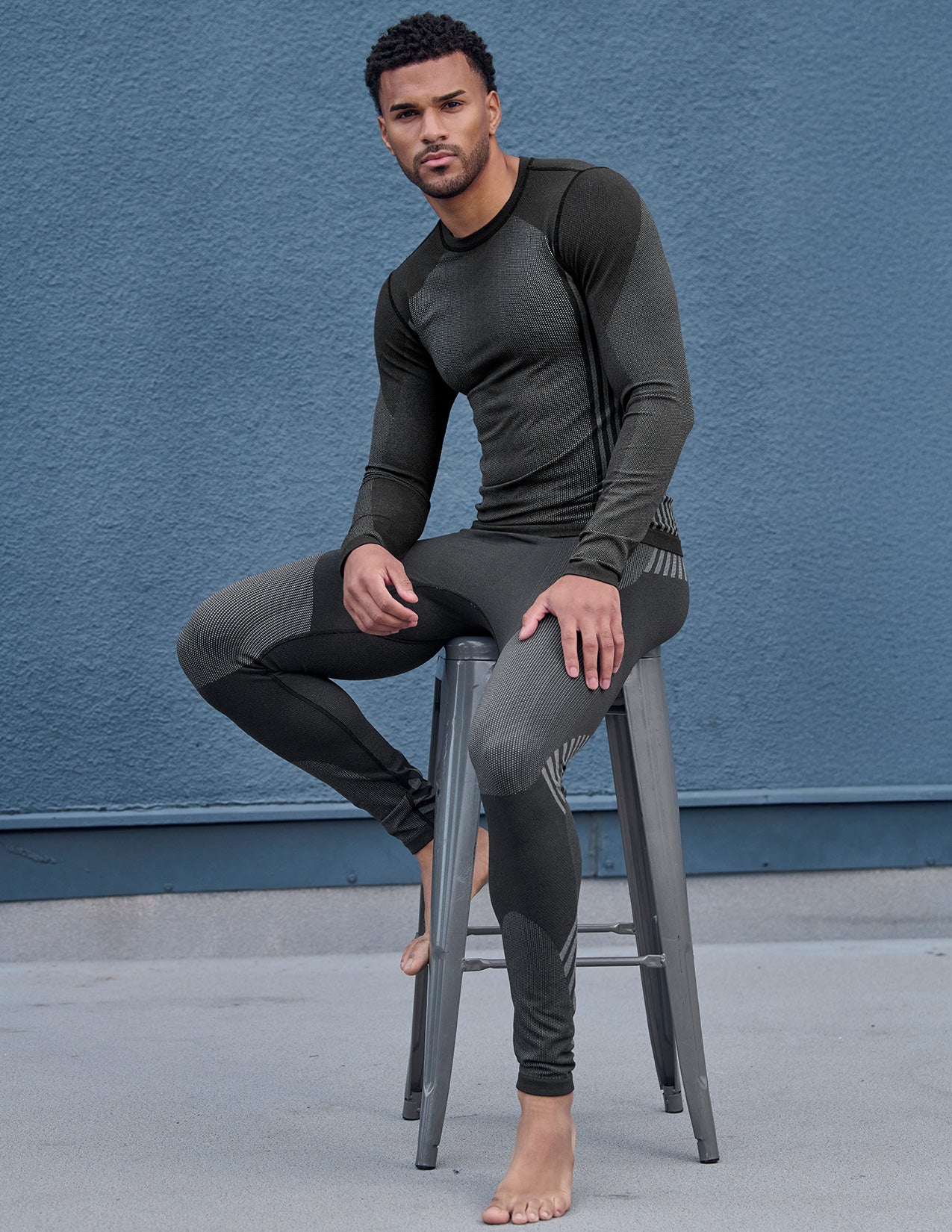 Man wearing a black thermal pants and longsleeve sitting on a stool against a blue wall.