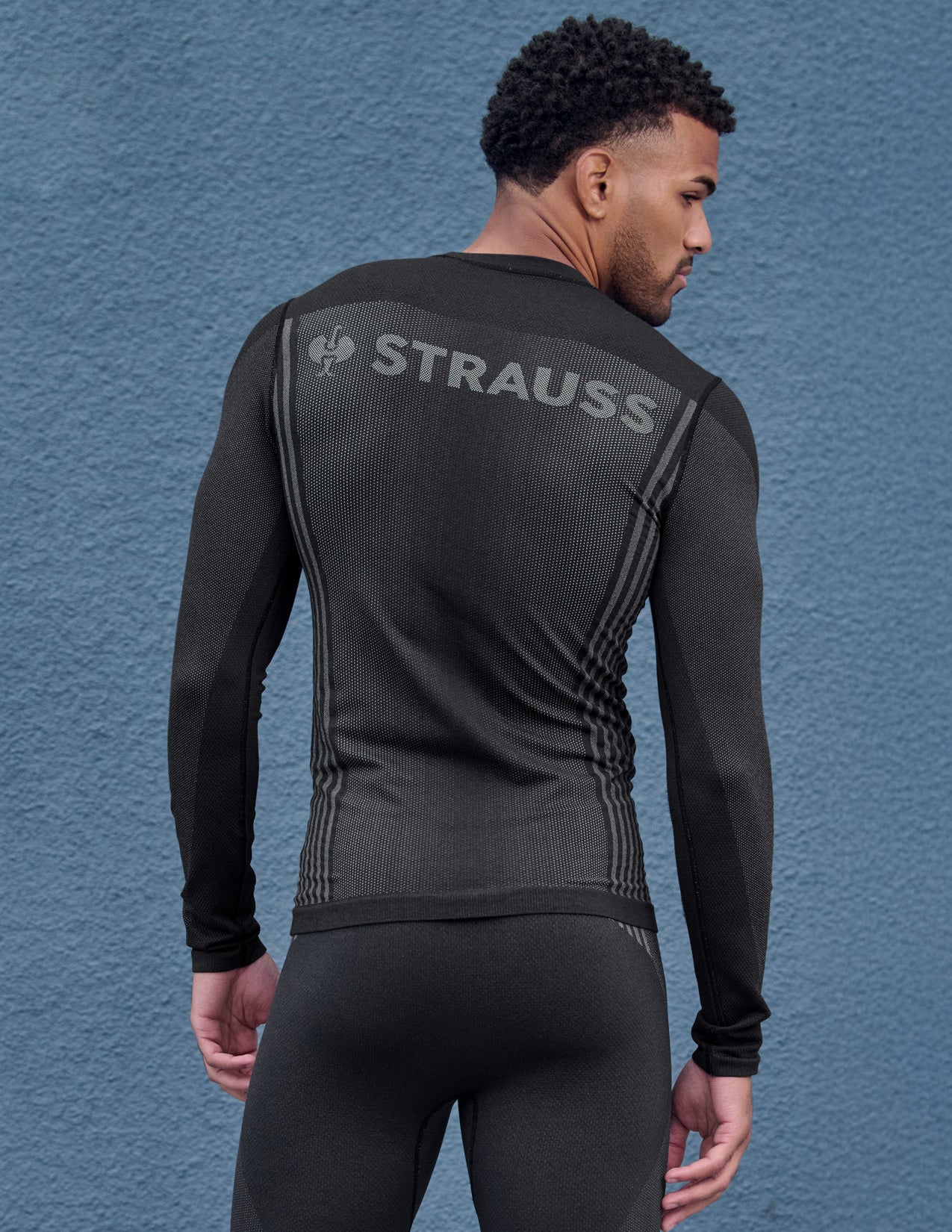 Man wearing a black thermal outfit with 'Strauss' branding on the back against a blue textured wall.