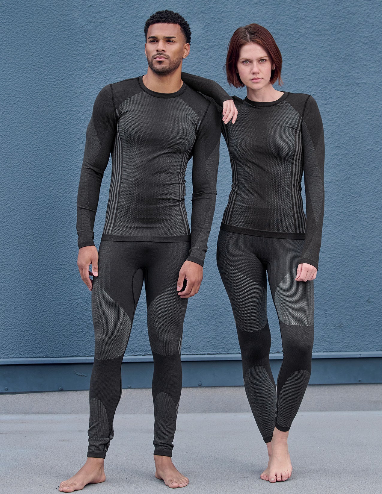 Two people wearing black thermal pants and longsleeves standing against a blue wall.