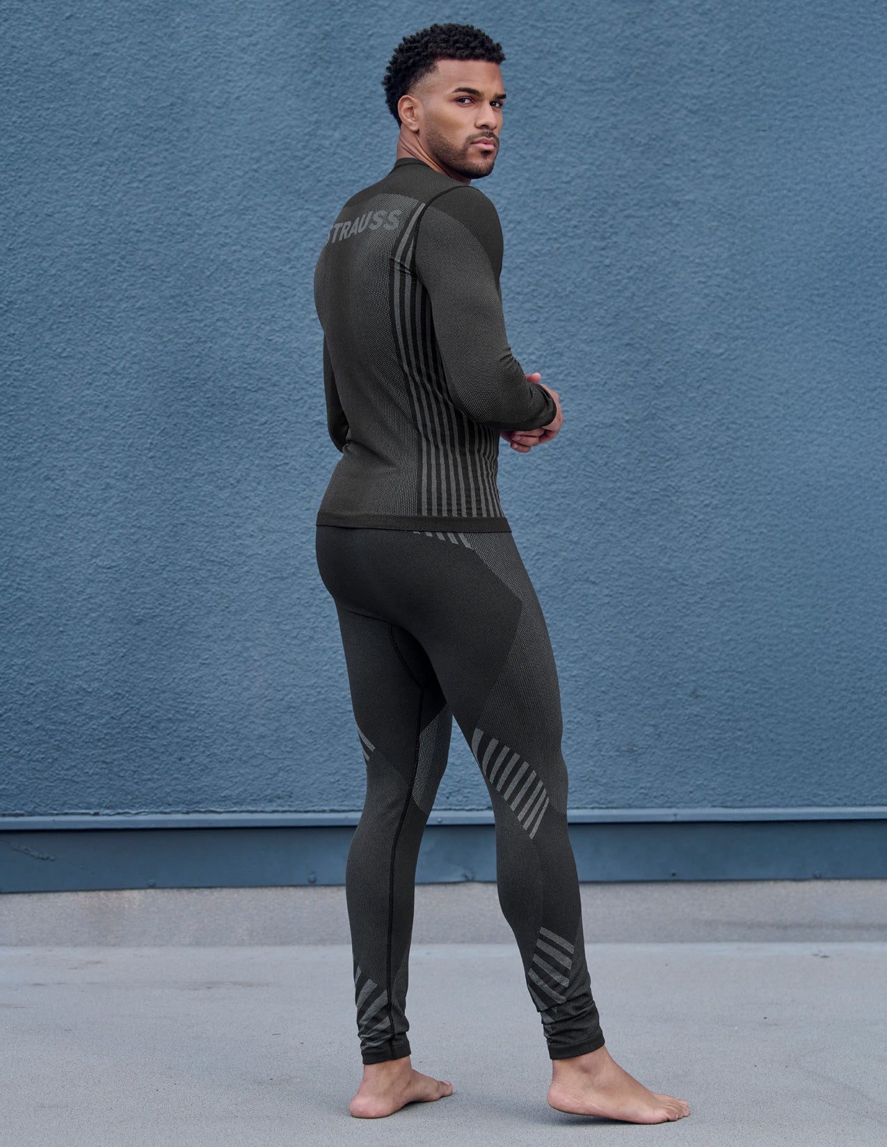 Man wearing a black thermal pants and longsleeve with STRAUSS branding against a blue wall.