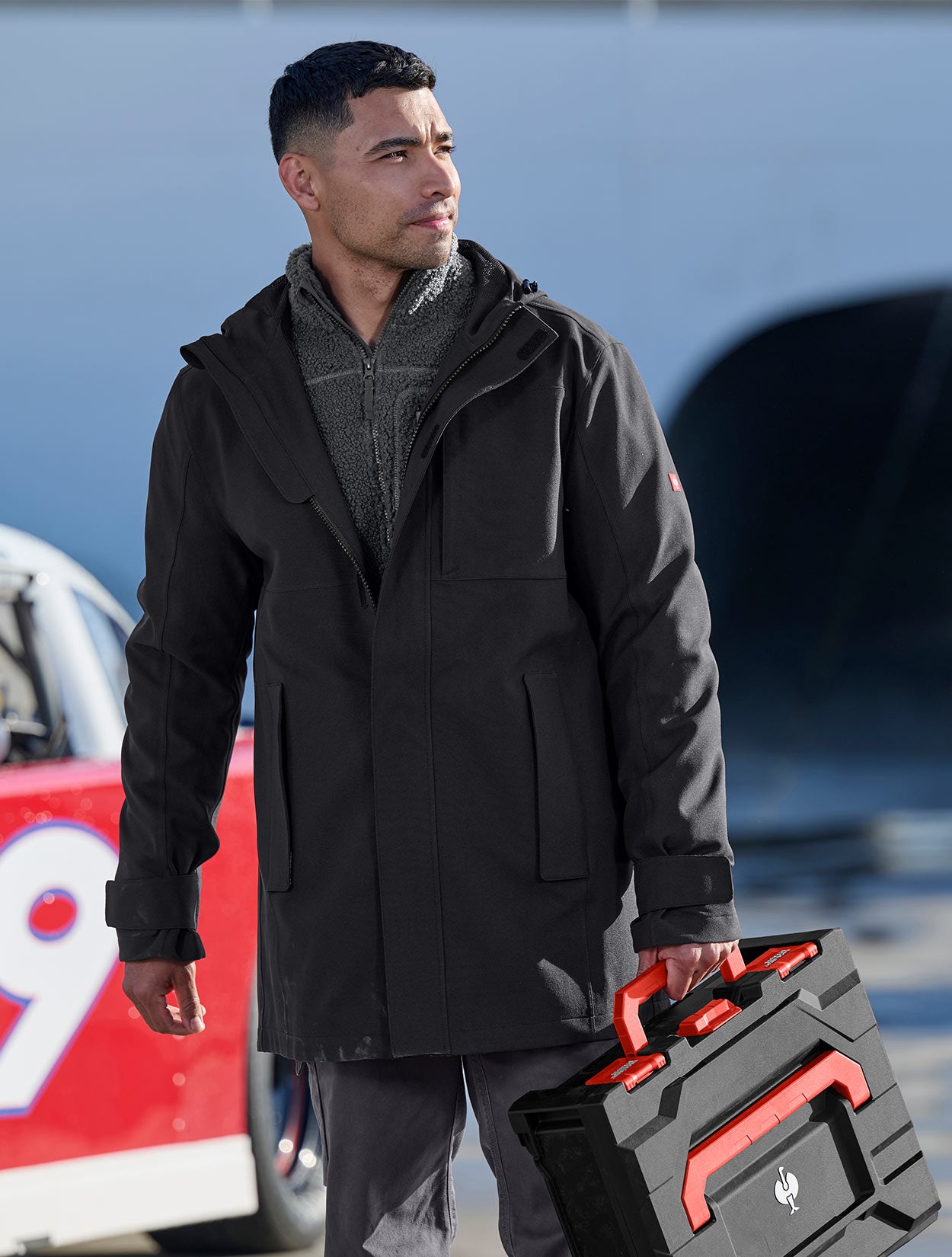Man in a black coat holding a red toolbox with a blurred background