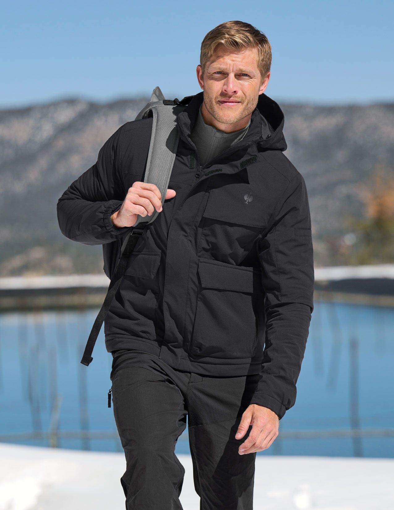 Man wearing a black jacket holding a backpack with mountains in the background