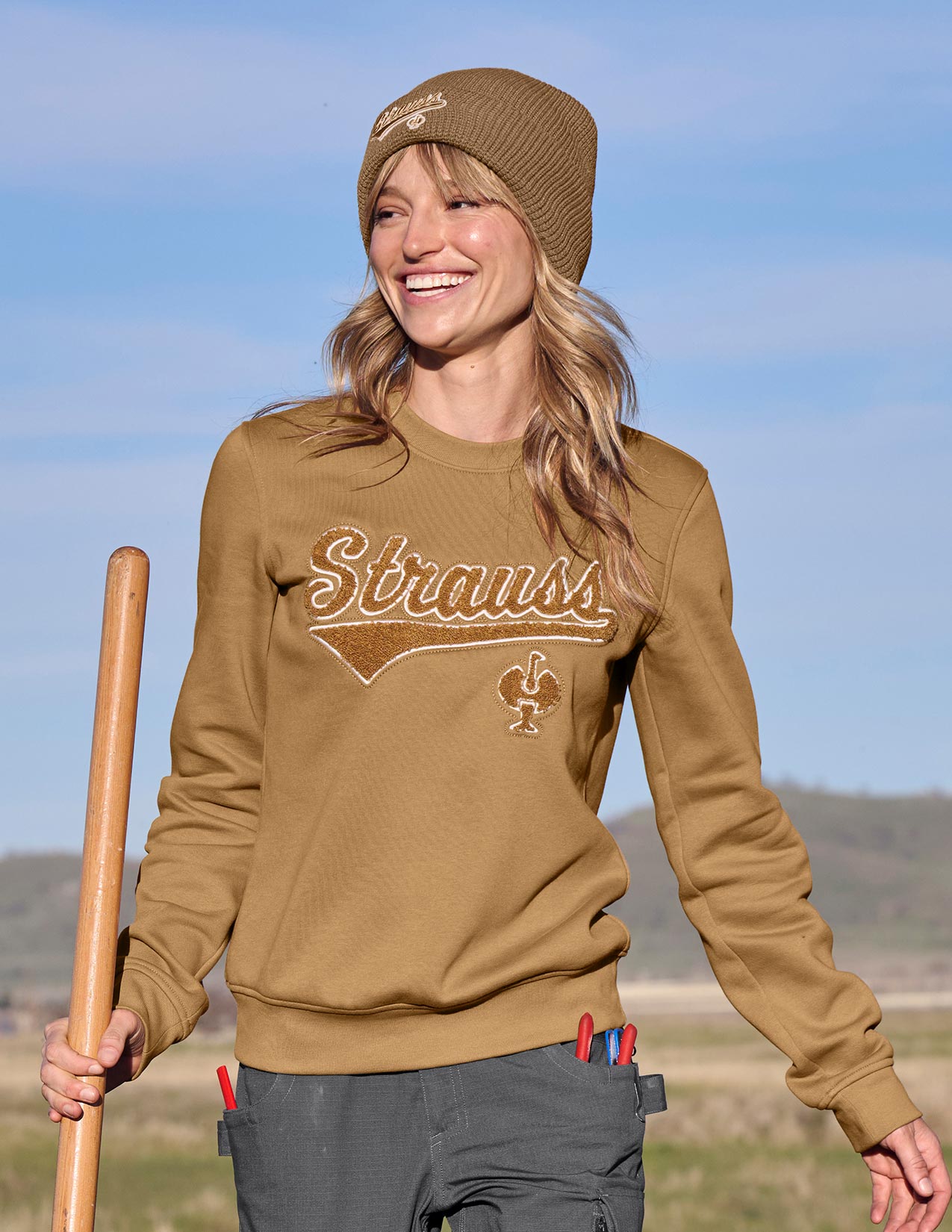 Woman wearing a brown sweatshirt with 'Strauss' logo, holding a wooden tool against a blue sky background.