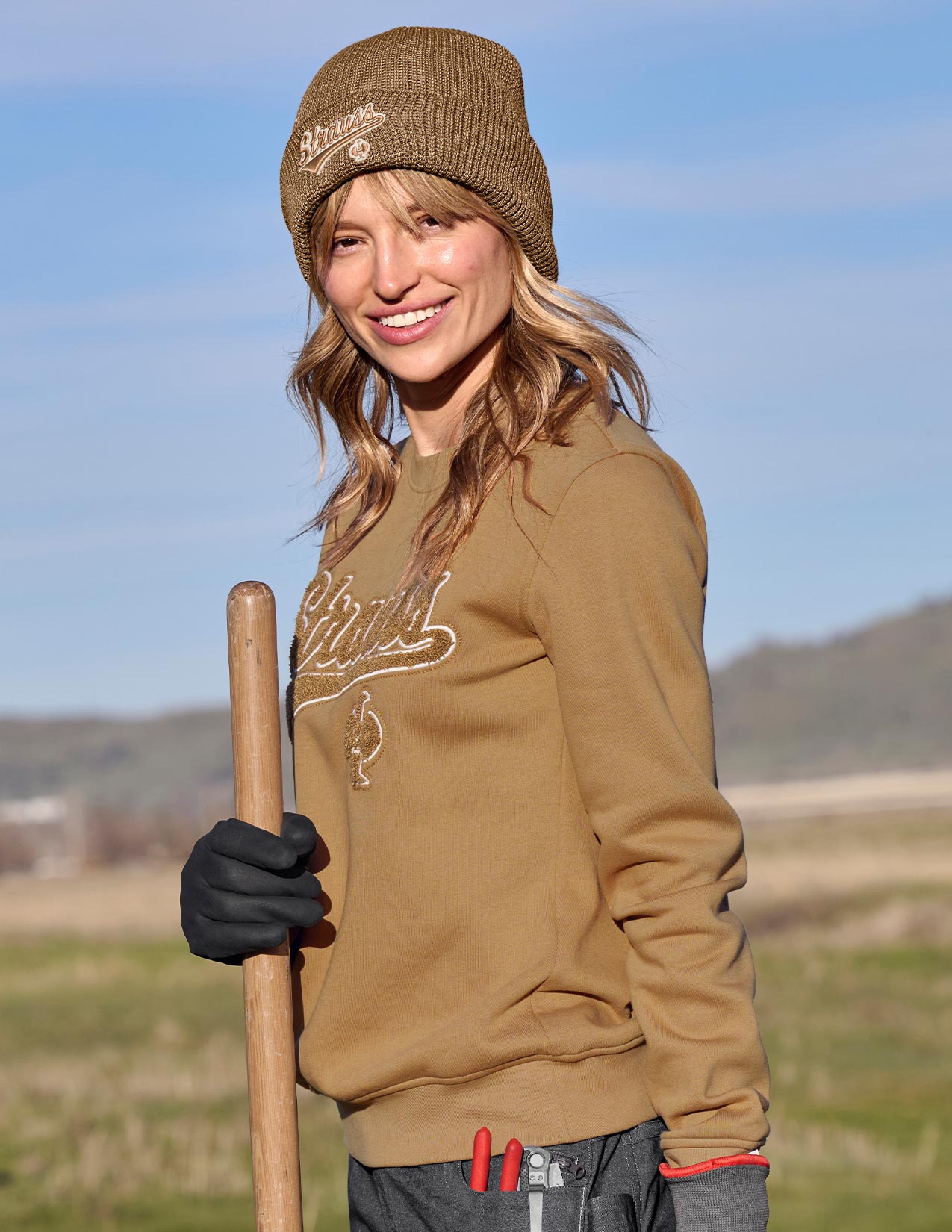 Woman wearing a brown sweatshirt and beanie holding a shovel outdoors.