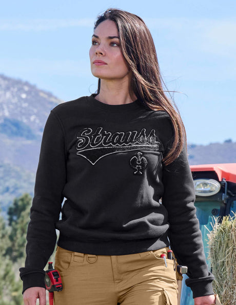 Woman wearing a black sweatshirt with 'Strauss' text, standing outdoors with mountains in the background.
